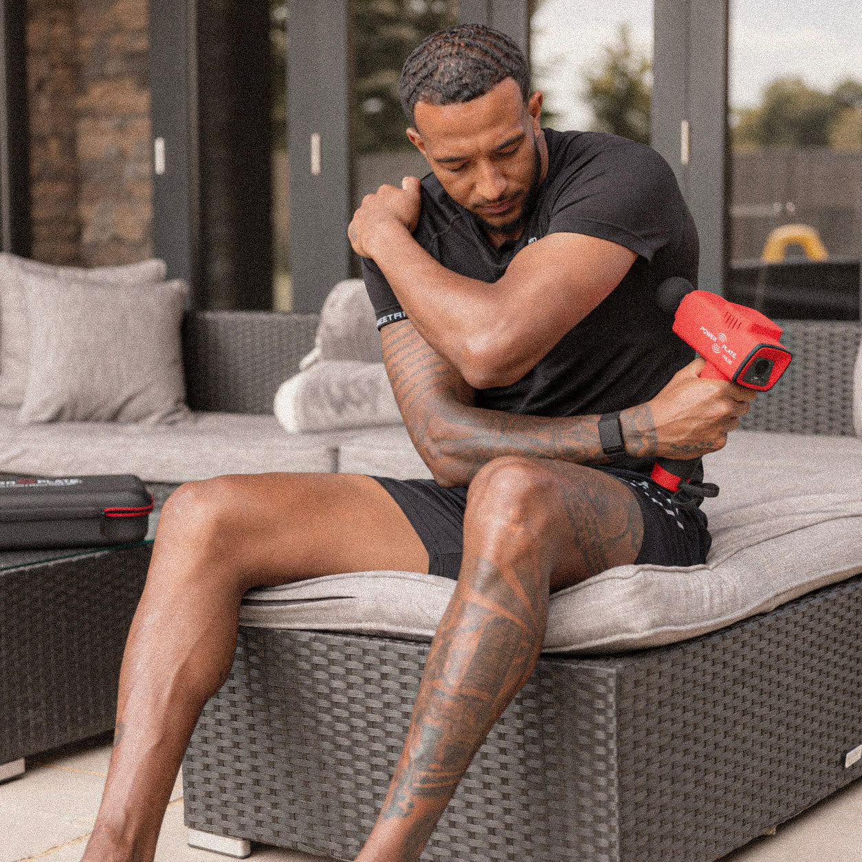 Man seated on a sofa using a red Power Plate Pulse on his upper arm.